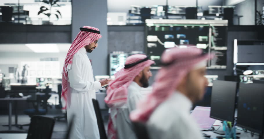 Arab Information and Cybersecurity Specialist Walking in the Office, Sitting Down to Work on His Desktop Computer. Young Middle Eastern Software Engineer Wearing a Traditional White Robe