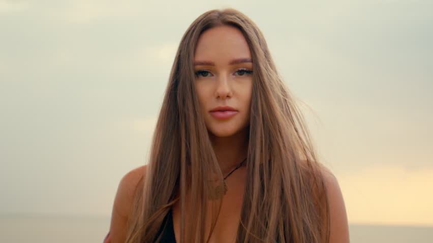 a beautiful girl with long straight hair stands on the beach and looks at camera