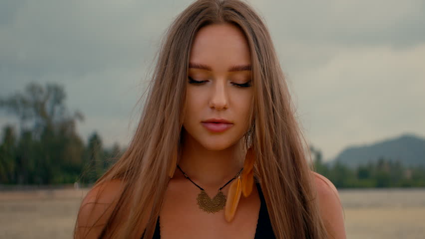 a beautiful girl with long straight hair stands on the beach and looks at camera