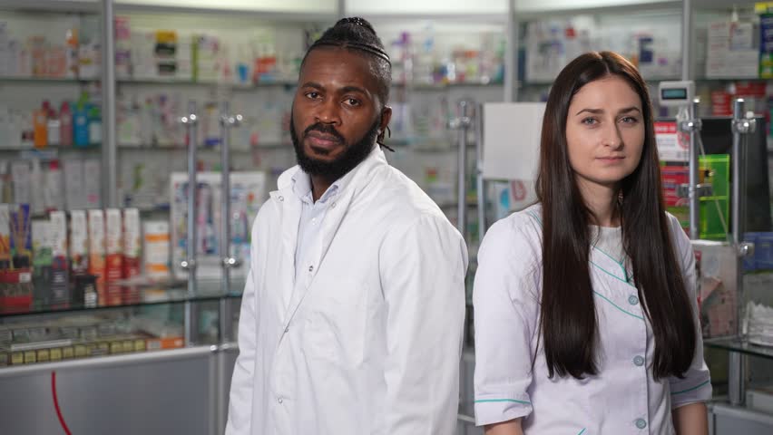 In this 4K high-quality footage, a black middle-aged male and young female pharmacy professionals smile for a portrait. The video highlights the work of drugstore staff in healthcare and medicine  | Shutterstock HD Video #1102170793