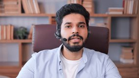 Successful Indian young man in headset having business video chat on laptop in office. Businessman talking to web camera holding video conference call with clients online, support call center web cam.