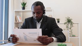 Motivated african employee in headset analysing market growth using charts and graphs during video meeting at office. Handsome man using wireless laptop for online conversation with coworkers.