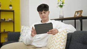 Young hispanic man watching video on touchpad sitting on sofa at home