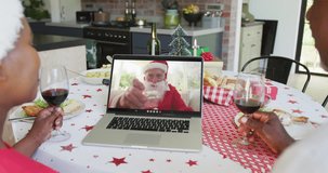 African american couple with wine using laptop for christmas video call with happy santa on screen. christmas, festivity and communication technology.
