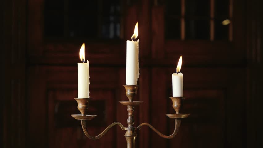 A rusty three-armed candlestick holdning three burning candles with the wax sliding down the sides. The out of focus background is an old rustic wood cabinet. Royalty-Free Stock Footage #1102178143