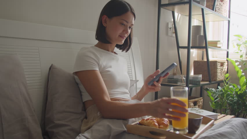 Young woman having breakfast in bed, drinking orange juice and using mobile phone in morning at home