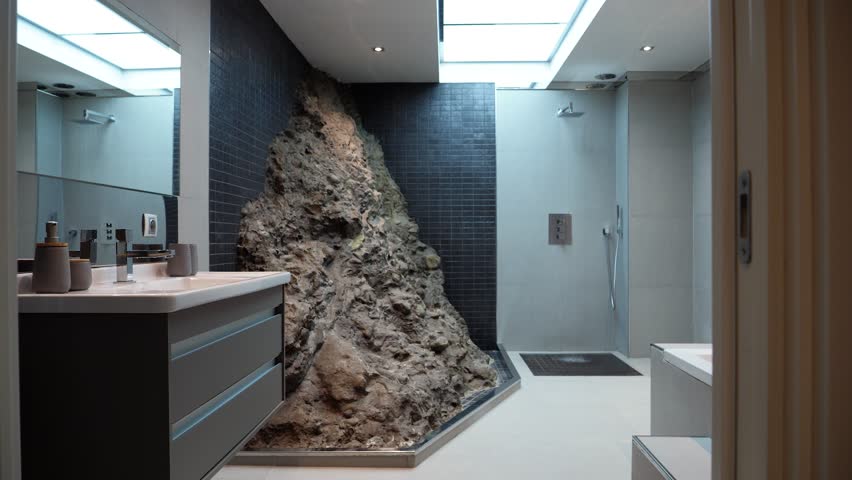 a modern and impressive bathroom with a rock wall in the center, a modern sink with mirror on the left and a bathtub on the right, a shower at the back, a skylight on the ceiling