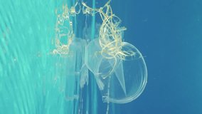 Vertical video, Close-up of plastic debris floating under surface in blue water, Slow motion. Disposable plastic cups and other trash littering oceans of planet.  
