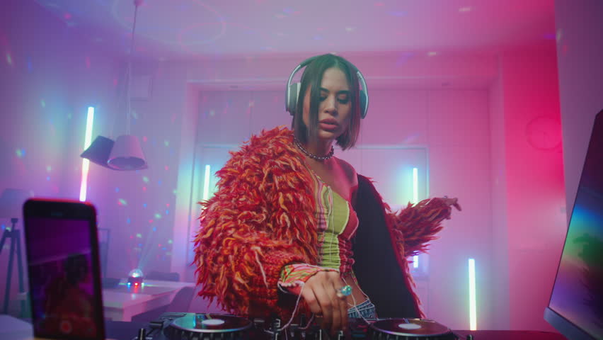 Stylish young woman in headphones listening techno music dancing create techno podcast using turntables at home, joyful female streaming dj set in room illuminated with colorful glowing neon lights