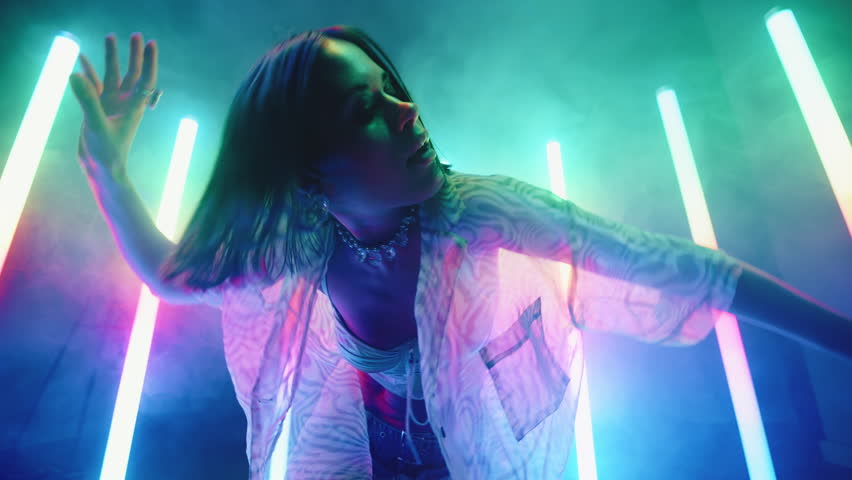 Stylish young woman dance with her hands backlit with colorful neon light lamps, female in trendy outfit dancing in night club, creative performer, haze and glowing tube lights, slow motion wide shot