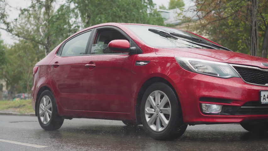Stylish red car drives on asphalt road in city in heavy rainy summer weather. Modern automobile moves on street against old buildings under pouring rain closeup