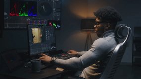 African American film maker or colorist does color grading on computer in studio. Big screen with RGB colour correction graphic bar on the wall. Action film footage displayed on monitor. Dolly shot.