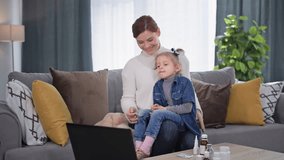 adorable caring female parent giving cold medicine to little daughter during online doctor consultation sitting on sofa, child licks tongue after taking cough syrup