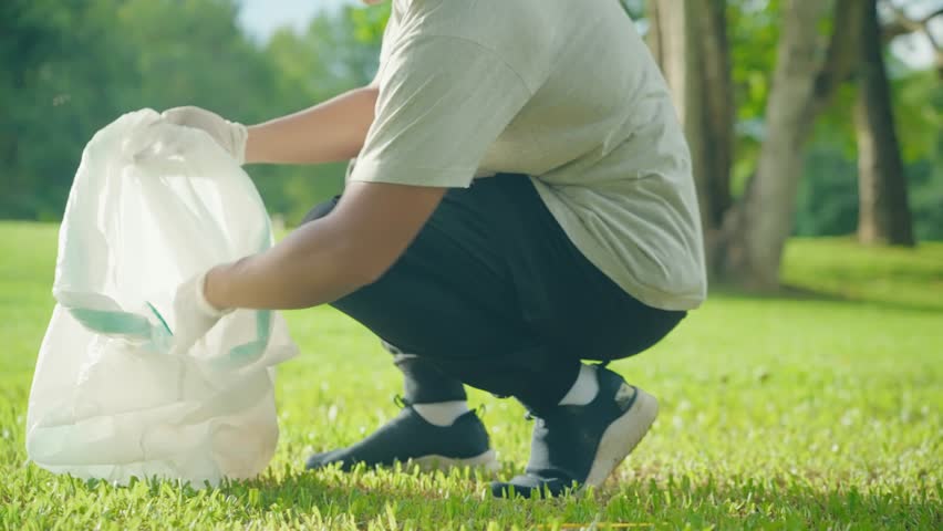 Closeup shot Volunteer's glove hands sitting, collects in bag a plastic trash on grass area inside the public park during the sunny day. Plastic pollution and environmental problem. Greening  planet Royalty-Free Stock Footage #1102223119