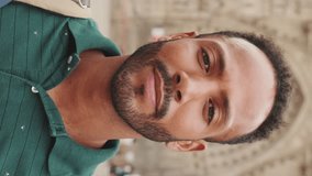 VERTICAL VIDEO, Close-up of young man tourist with backpack on his shoulder, looking at the camera and smiling while standing on the square of the old city