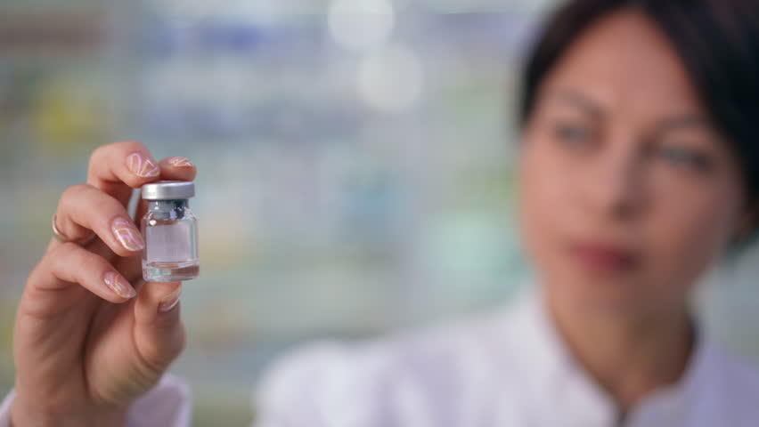 Rack focus from vaccine jar in female hand to concentrated serious woman examining contradictions. Portrait of focused Caucasian pharmacist choosing treatment standing in drugstore Royalty-Free Stock Footage #1102224431