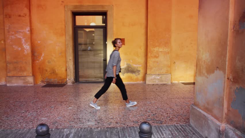 Young attractive female blonde model wearing striped t-shirt and dark pants walking around in the streets of Bologna, Italy, in front of bright orange walls of ancient European buildings Royalty-Free Stock Footage #1102224663