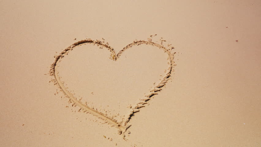 Close-up of a heart sign written on sand being washed away by the sea waves reaching the shore of a beach