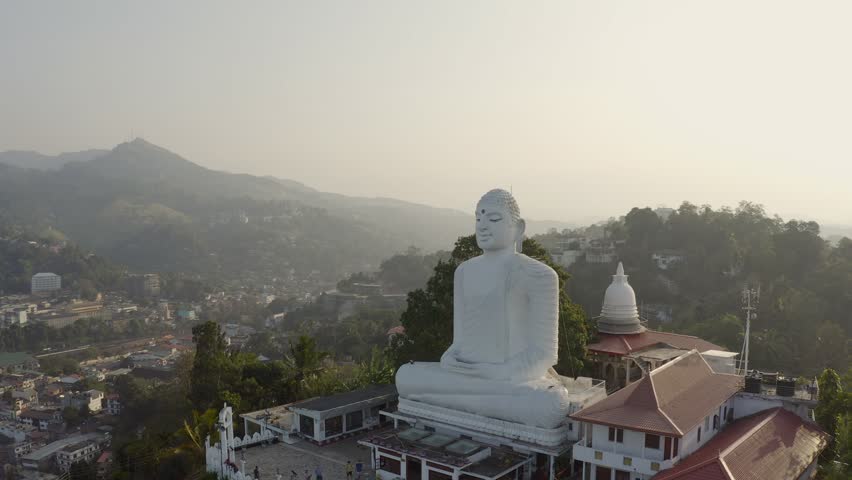 Drone point of view of big white sitting Buddha overlooking the green lush hills around. Aerial view of buddhist temple in Kandy, Sri Lanka at sunset