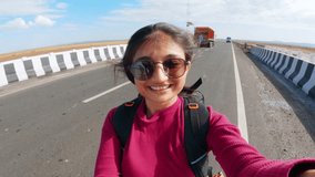 Portrait of Beautiful Indian girl traveler wearing sunglasses and enjoying bike ride while sitting on backseat. Happy Female taking selfie video from moving vehicle in Khavda, Kutch, India. 