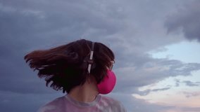 Young caucasian brunette woman sunglasses, pink mask and headphones dancing, listening music on the background of cloudy sky. High quality 4k video footage