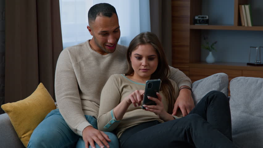 Multiracial couple customers relax on sofa use mobile phone at home diverse woman wife and man husband talk make order online delivery buy internet goods shopping in e-commerce app look at cellphone Royalty-Free Stock Footage #1102240991