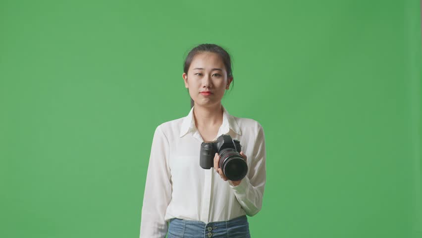 Asian Photographer Using A Camera Taking Pictures And Smiling Touching Her Chest While Standing On Green Screen Background In The Studio
