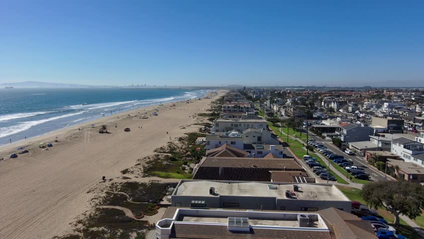 backwards aerial footage flying along the coastline at Rosie's Dog Beach with people walking along the beach, homes and cars on the street with blue ocean water and waves rolling in in Long Beach