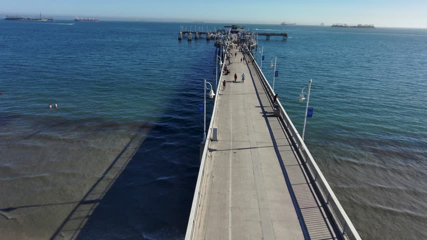 aerial footage of a long concrete pier over the vast deep blue ocean water with people walking along the pier and curved light posts along the pier at Belmont Veterans Memorial Pier in Long Beach