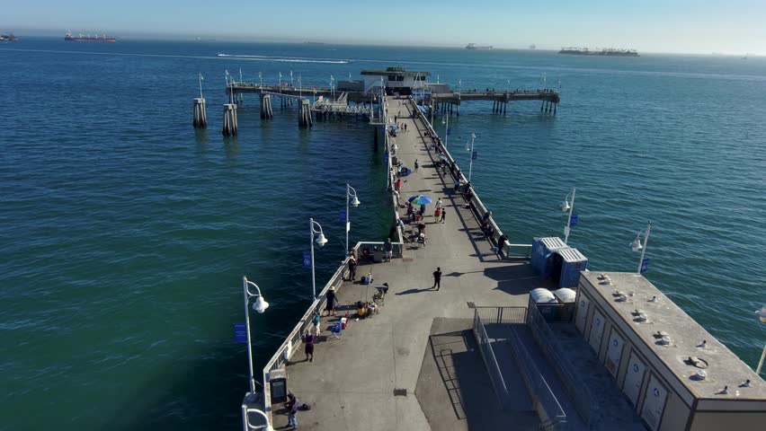 aerial footage of a long concrete pier over the vast deep blue ocean water with people walking along the pier and curved light posts along the pier at Belmont Veterans Memorial Pier in Long Beach