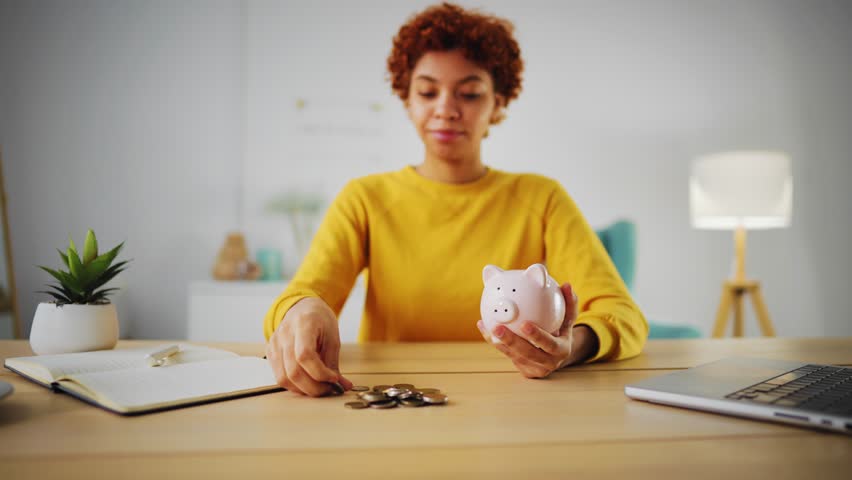 Portrait of curly smiling african american woman wearing yellow sweatshirt holding pink piggy bank in hands and putting coins inside it. She sits at desk, workplace. Saving money self finance concept. Royalty-Free Stock Footage #1102252911