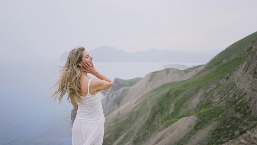Young woman blonde in white dress touches long loose hair waving on wind posing for camera and standing on rocky cliff edge against mountains and blurred sea close backside view