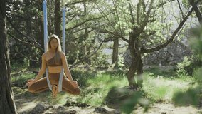 Young woman in slim bodysuit practice aero-yoga exercise, meditate in lotus pose hanging on ropes on tree branch against green bushes in bright backlit, tracking shot - Powered by Shutterstock - Get 15% off with code: PIKWIZARD15
