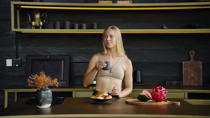 Young blond woman having breakfast in her kitchen at home alone, single girl student enjoy her coffee break eating bakery and drinking coffee, concept of self-care self caring, being independent