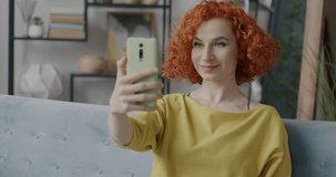 Portrait of happy young woman making online video call with smartphone speaking and waving hand in apartment. Communication and people concept.