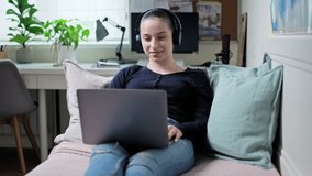 Teen girl in headphones looking at laptop screen, lying on couch at home