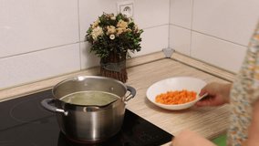Video of cropped woman hands putting, adding carrot vegetable diced cubes as ingredient for soup in boiling water in steel pan on induction cooker in kitchen. Cooking healthy meat soup, 4 k resolution