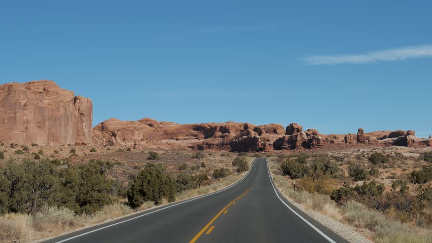 Driving vehicle on empty smooth highway going into distance to red mountain rocks in canyon landscape in dried hot desert Arches national park. Highway with black asphalt and orange road markings.