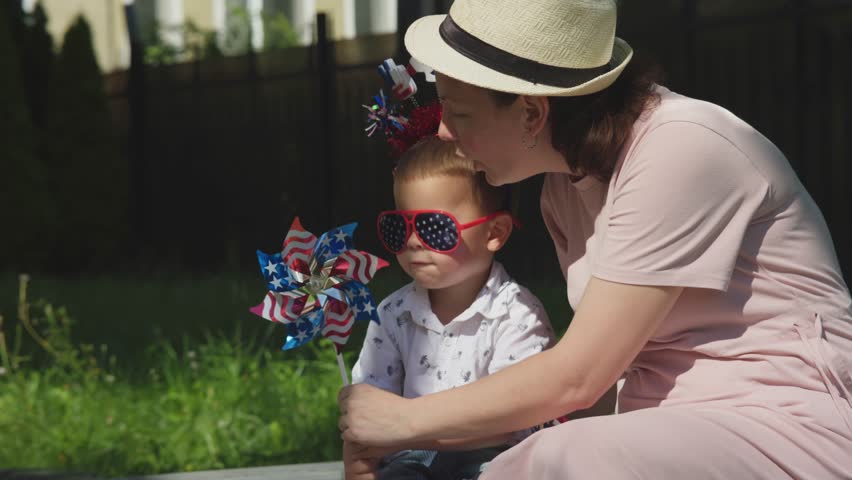 Happy american family concept mother and son sitting green grass on 4th July, mom teaching boy kid blow stripes and stars windmill. smiling happy Independence holiday outdoors of backyard  Royalty-Free Stock Footage #1102285565