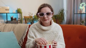 Excited young woman sitting on sofa eating popcorn and watching interesting tv serial, sport game, film, online social media movie content at home. Girl in 3D glasses enjoying domestic entertainment