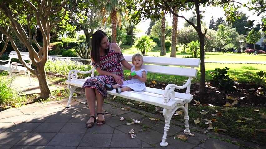 Mom and little girl are sitting on a white bench in a green park