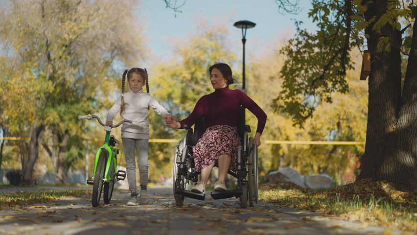 Loving mom and little daughter with long braids walk in park. Woman with spinal cord injury and preschooler hold hands tenderly and smile in joy of communication