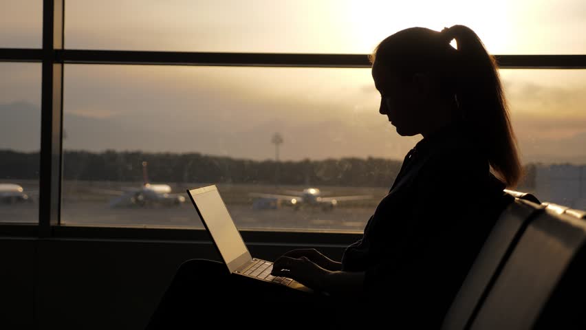 Busy woman works even while waiting for flight at airport lounge. Silhouette of passenger typing at laptop, sitting against window. Bright sunset light shine outside, blurred apron on background Royalty-Free Stock Footage #1102303295