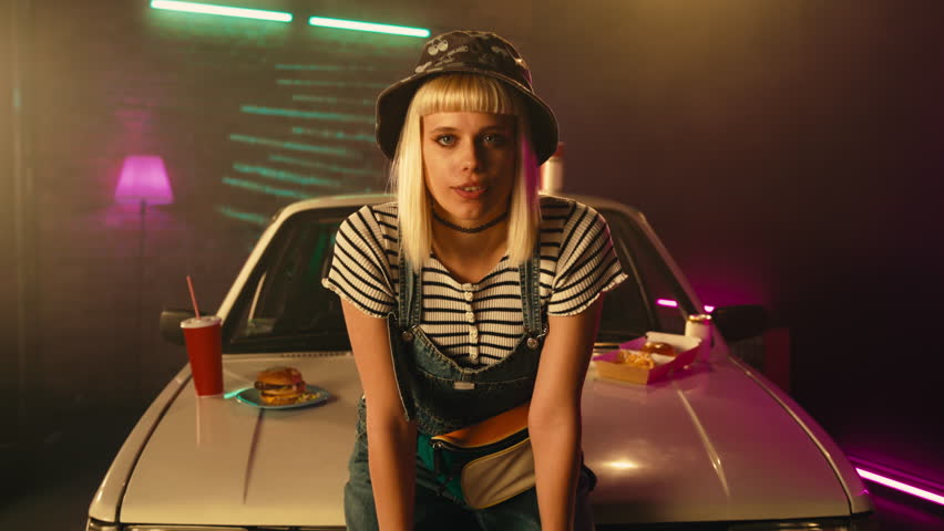 A vintage styled woman is relaxing on the hood of her car with fast food next to her.