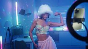 Creative female in trendy outfit recording video for social media, disco dance at home. Stylish woman in white hat dancing moves rhythmically listen music in room illuminated with colorful neon lights