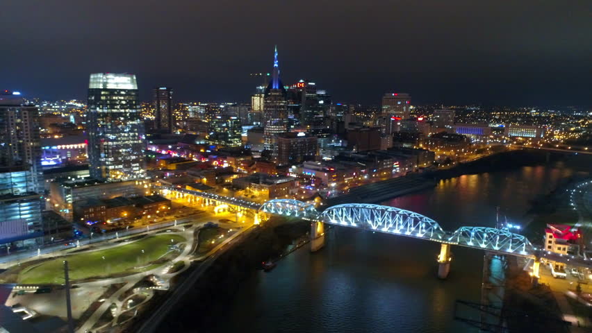 Aerial Panning Shot Of Illuminated John Seigenthaler Pedestrian Bridge By Modern Buildings In City Against Clear Sky At Night - Nashville, Tennessee Royalty-Free Stock Footage #1102310443