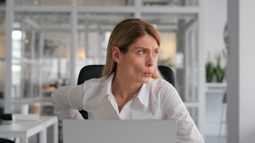 Woman working on laptop in office is experiencing back pain. Prolonged sitting and poor posture are common causes of back pain in office workers. Need take breaks, stretch, and maintain good posture. | Shutterstock HD Video #1102316773