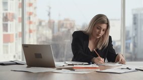 A young business woman checks the company's financial statements while sitting at a desk in her office. A blonde woman in a business suit makes notes on various documents that are spread out on the