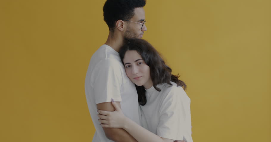 Slow motion portrait of happy young Caucasian woman hugging African American boyfriend and smiling looking at camera on yellow background