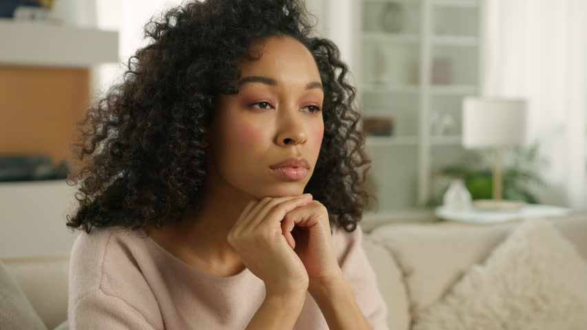 Close up face sad thoughtful young African American single woman looking away thinking of problems. Woman of color siting on sofa alone at modern home troubled with negative thought in slow motion 4K Royalty-Free Stock Footage #1102332887